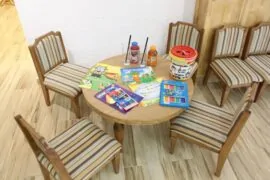 Детски кът - Малка маса с цветни детски книжки, играчки и напитки, разположена на уютни дървени столчета.