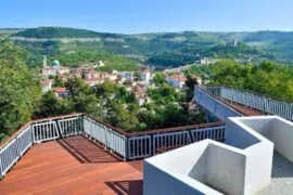 Панорамна гледка от наблюдателна площадка с дървени настилки, предлагаща изглед към зелени хълмове и открито пространство с жилищни сгради.