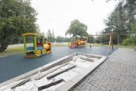 Площадка с люлки и влакче, разположена на меко покритие, подходящо за деца. Първоначалните стелажи и дърветата в парка добавят зеленина на фона.