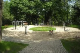 Площадка за фитнес на открито, с различни уреди за тренировки, разположена сред зеленина и пейки. Пътеките са покрити с каменни плочи и има обширно озеленяване.