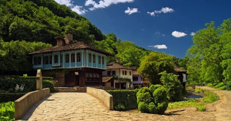 A shot of traditional houses surrounded by green bushes and trees, under a bright blue sky with clouds. The photo highlights the harmony between architecture and nature.