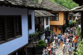 A group of visitors explore the architecture of the Etara Museum. Colorful houses with wooden sheds and roofs, surrounded by greenery, are visible.
