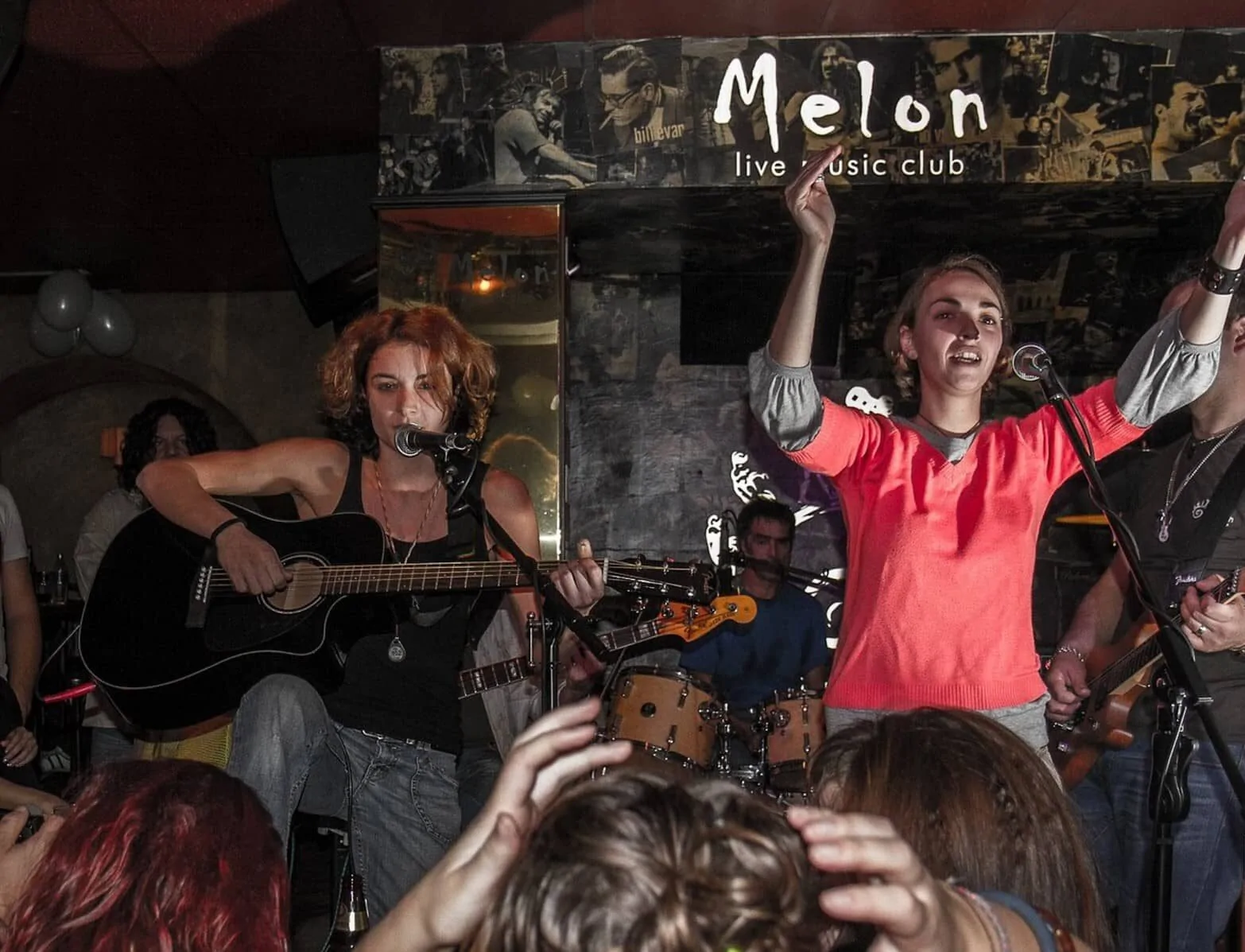 Melon Music Club - Live performance in a club with a lot of people. Two singers, one playing acoustic guitar. Lively atmosphere, people in the audience enjoying themselves.