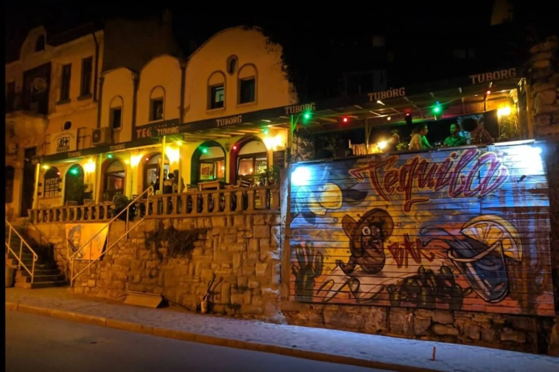Evening view of Tequila Bar Funky Monkey. Showing the more illuminated part of the establishment with colored lamps. There is graffiti related to tequila on the facade.
