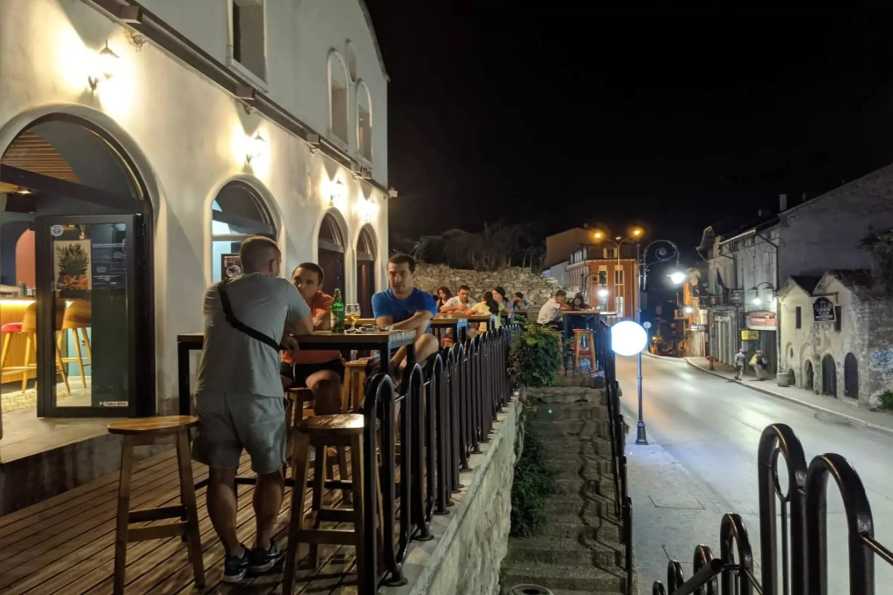 A group of people sitting outdoors at Tequila Bar. The illuminated terrace of the establishment with bouquets of flowers. A view of the street with passersby and decorations.