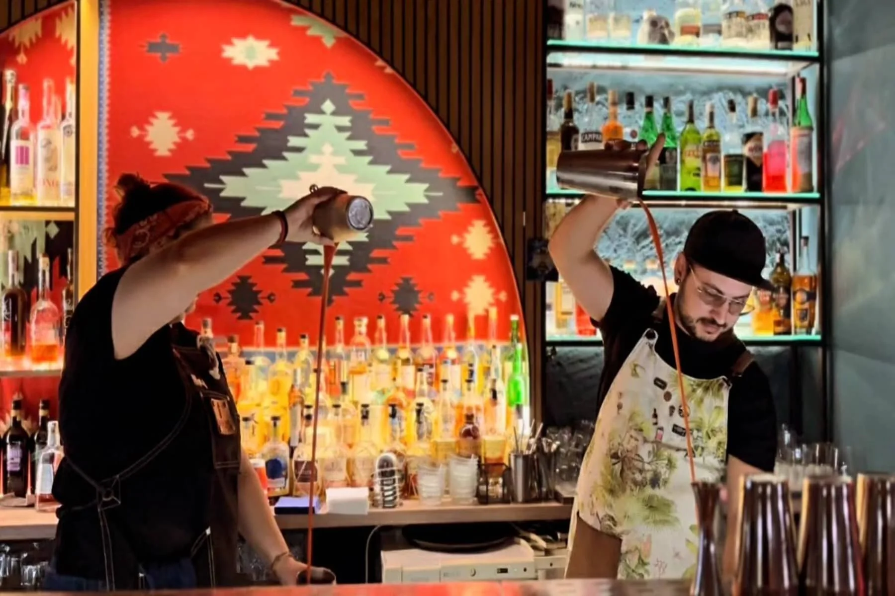 Two bartenders working behind a bar. Different techniques of mixing and preparing drinks. On a given background, shelves with a variety of alcoholic beverages and a colorful back wall are visible.