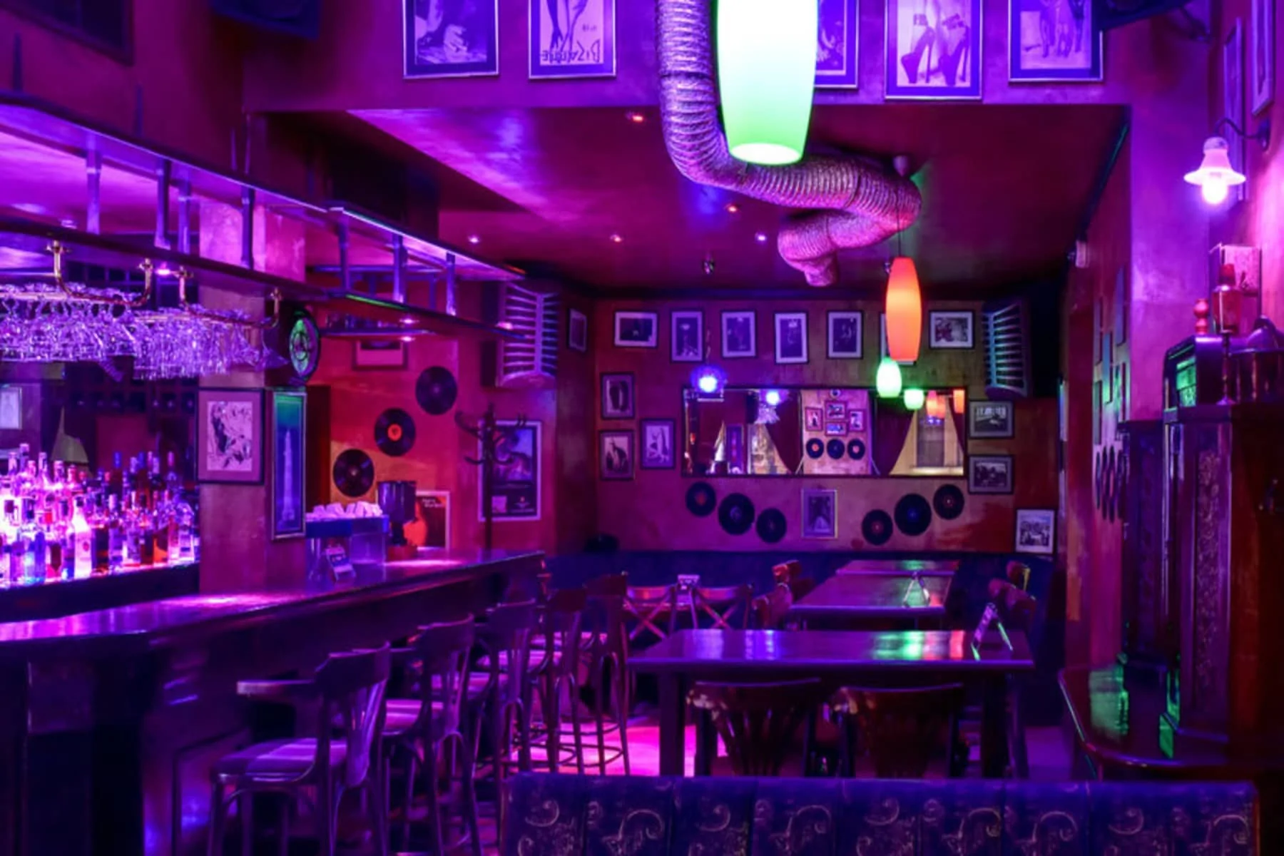 Interior of the second lounge of the bar, with bright lighting in purple and green. The bar has a classic wooden design, with many tables and chairs, and the walls are decorated with gramophone records and black and white photos, giving a retro atmosphere.