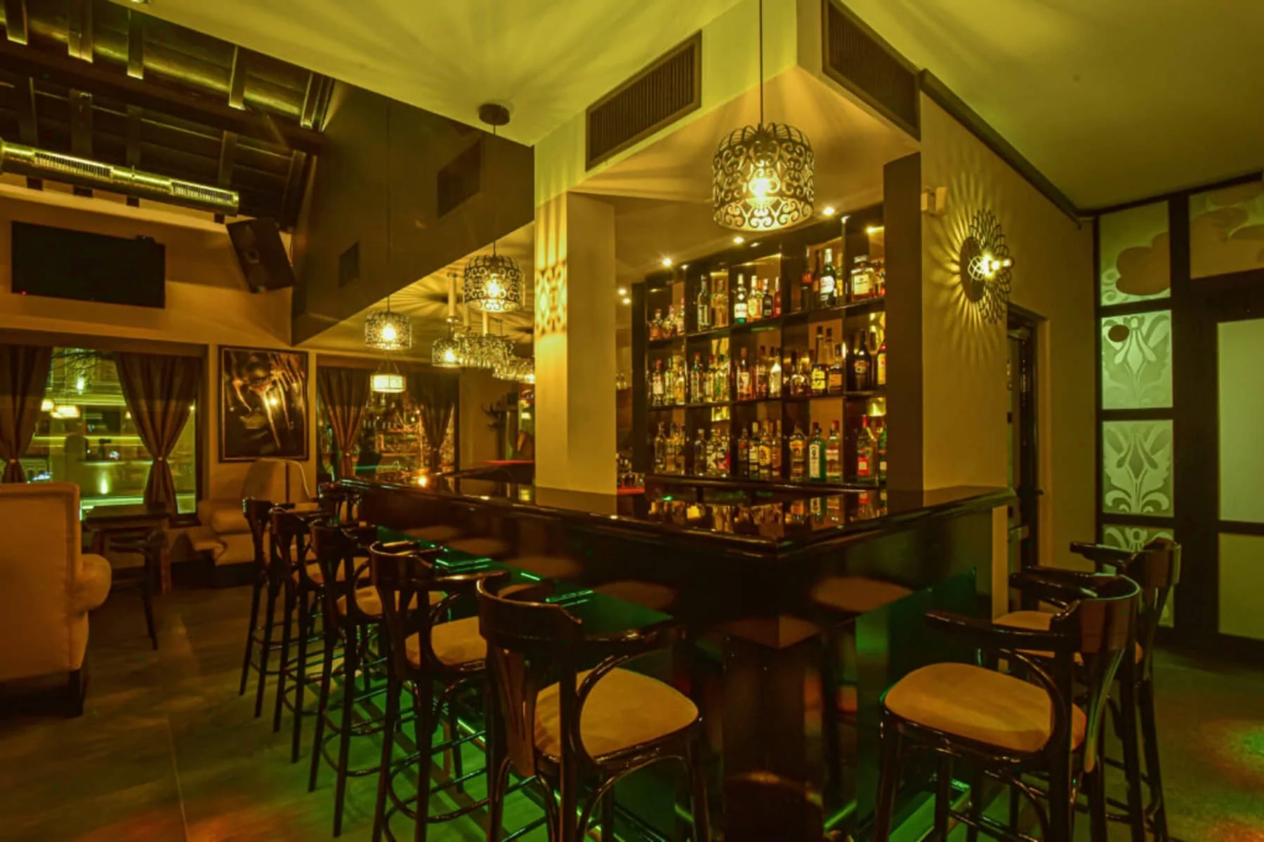 Bar interior Cozy bar interior with a dark wooden bar and a variety of drinks displayed on a shelf. Lighting in soft yellow and green tones, seating on solid bar stools. The walls are decorated with artistic elements and hanging photos.