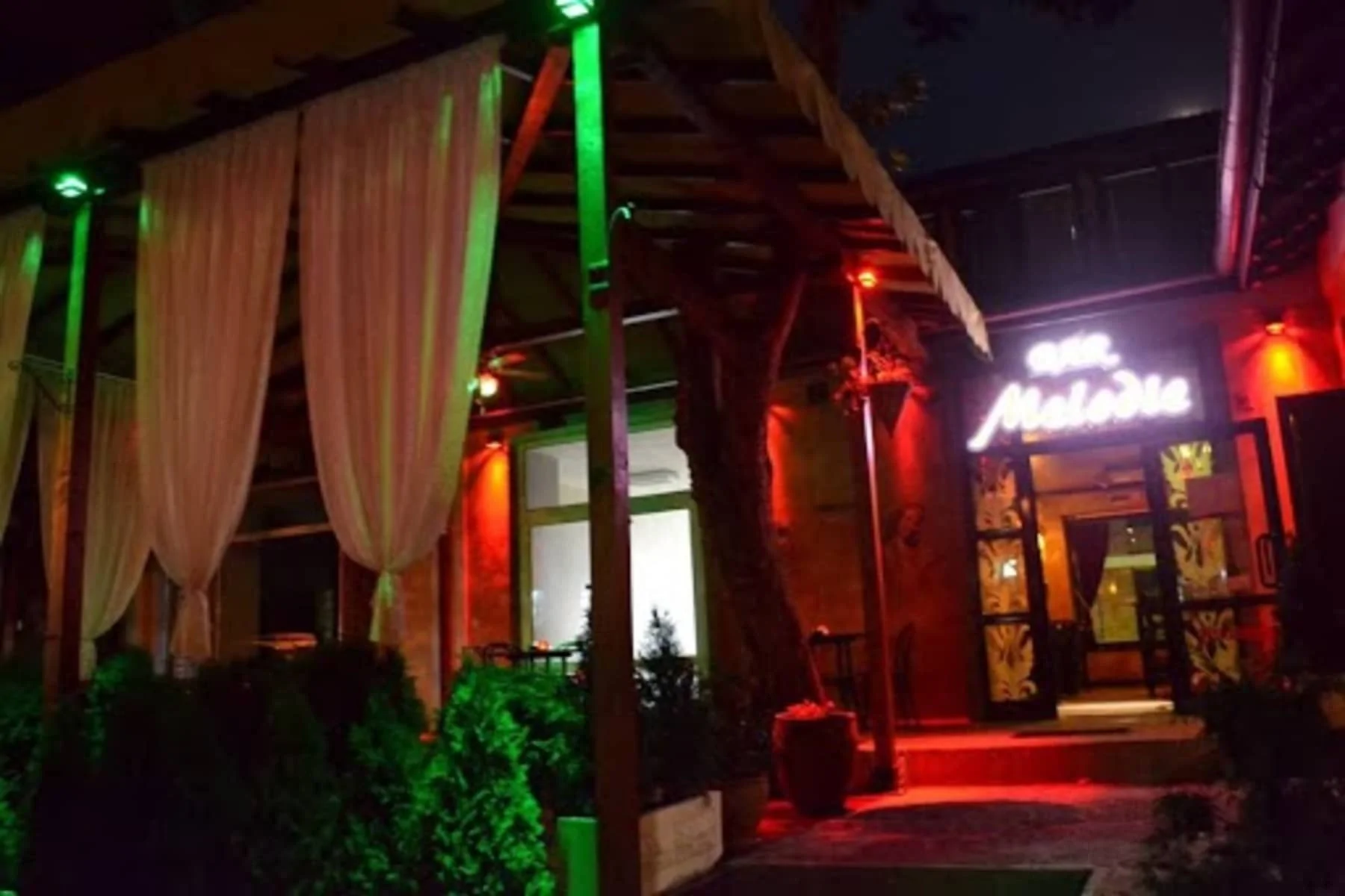The entrance to the Melodie bar with a contemporary sign and colorful lighting. The decoration includes white curtains and green bushes. The atmosphere at night is distinctly romantic, with bright red and green lights around the entrance.