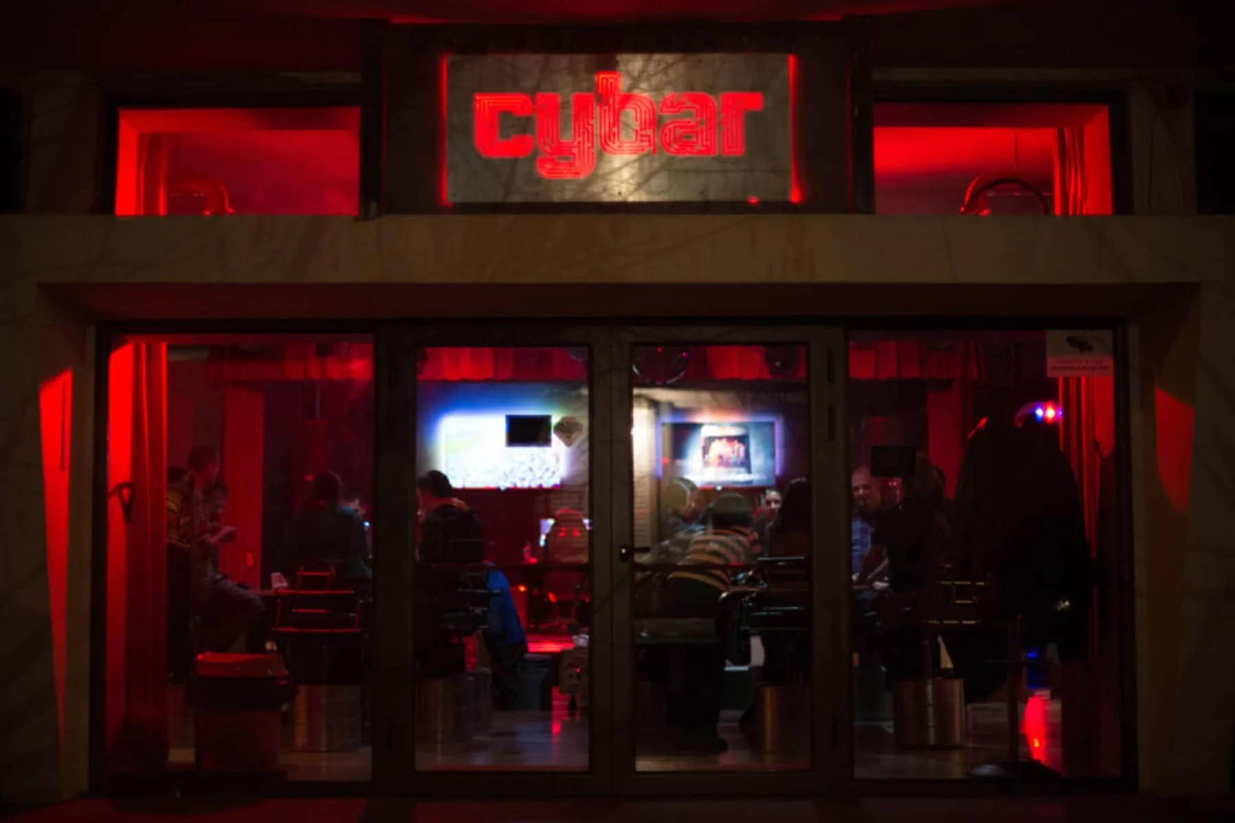 A view of the entrance to Cybar, lit in red tones. The windows show a busy interior with customers, and the neon sign with the Cybar logo is located on the facade.