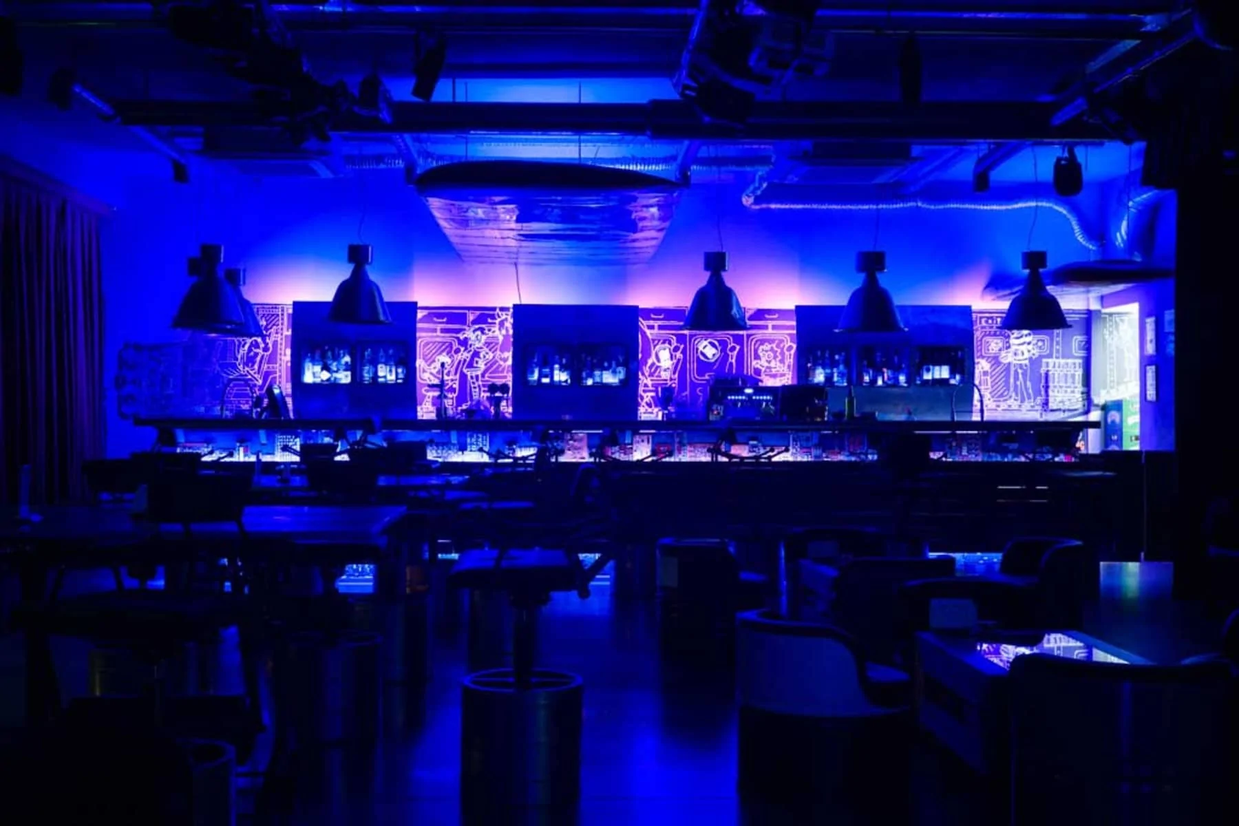 Cybar's interior, lit in blue tones. The bar has a prominent wall with graphic elements, and hanging lamps above it give the space a modern look. In the background, drinks can be seen behind glass display cases.