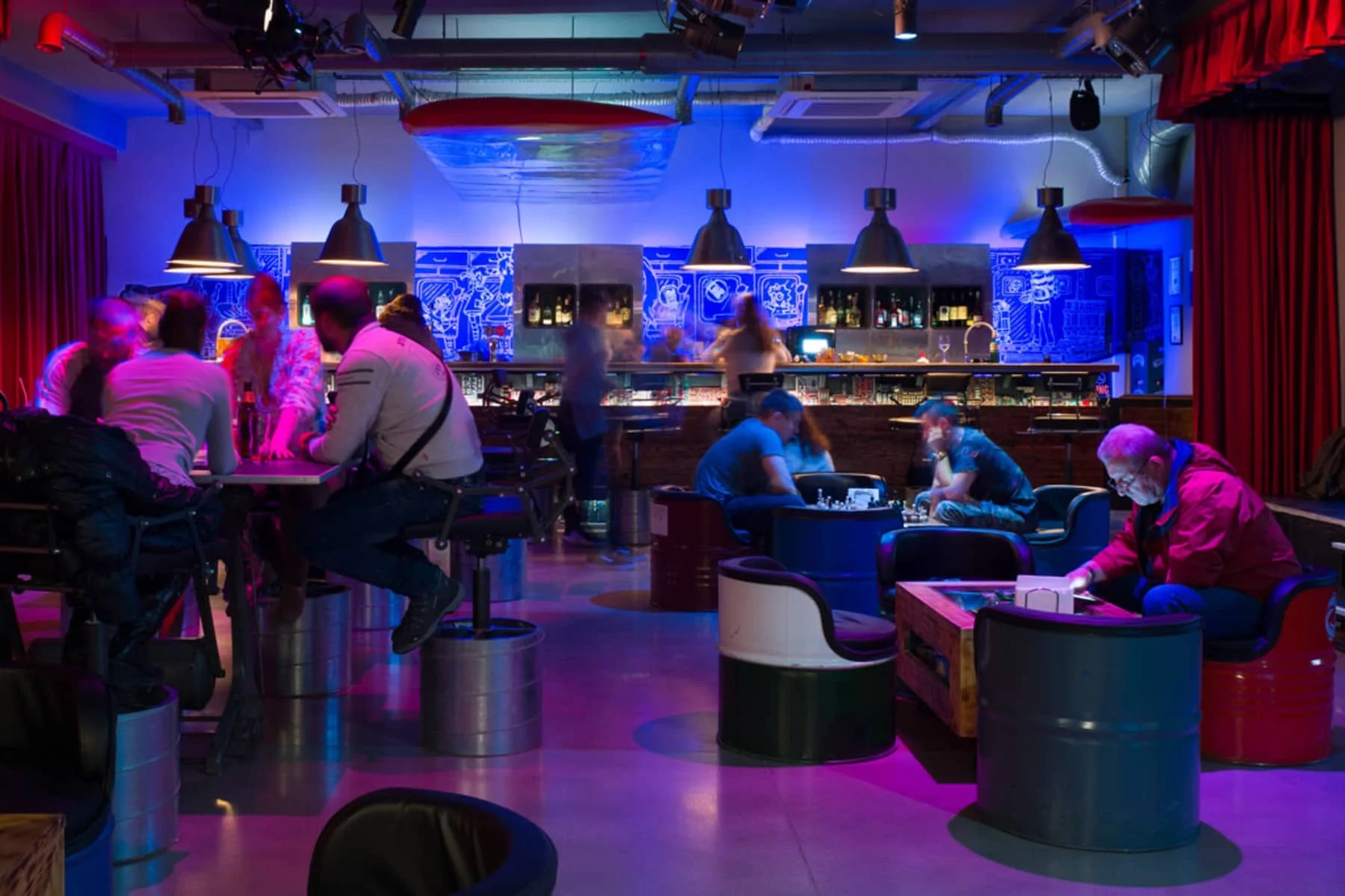 A back view of Cybar with a crowd of patrons. Chairs and tables made of various materials provide a cozy setting. Some customers play chess, while others chat, amidst shades of blue and purple lighting.