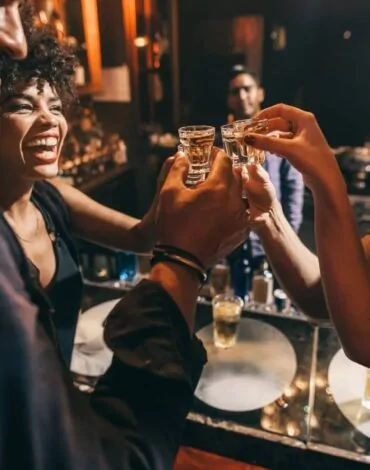 Група приятели по време на празненство в бар. Те вдигат тост с шотове алкохол. В центъра на кадъра се вижда усмихната жена с къдрава коса, която се радва на момента. На заден план се виждат мъже, които също се наслаждават на вечерта, с напитки на бара. Атмосферата е весела и празнична, с меки светлини около тях.
