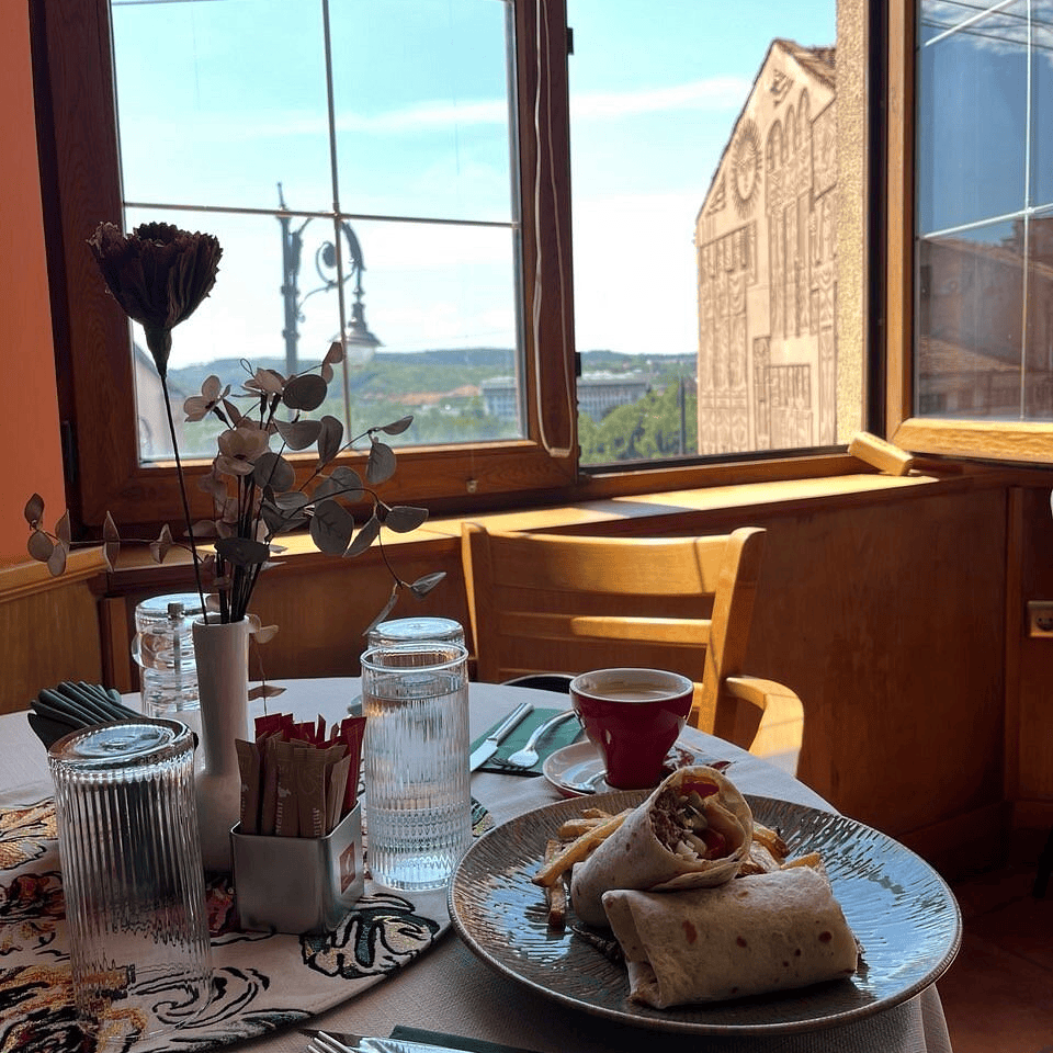 Restaurant interior with a window opening to a beautiful landscape. On the table there is a flower, a cup of coffee and a plate of burritos and fries. The atmosphere is cozy with wooden elements in the design.