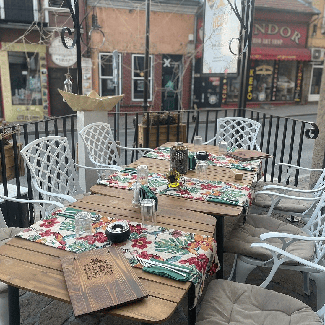 An outdoor terrace of a restaurant with tables covered with colorful tablecloths. The tables are arranged with glass glasses, salt shakers, and menus. Shops and characteristic architectural elements are visible in the background.