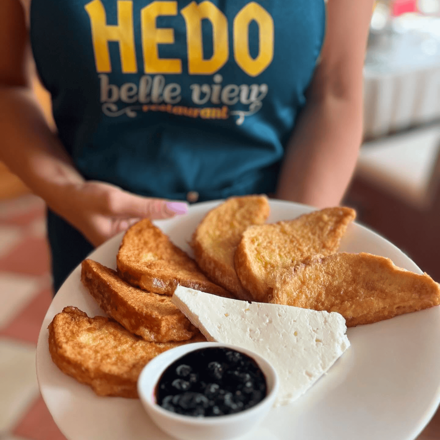 A plate of fried slices served with a slice of cheese and a bowl of cherry jam. In the foreground is a person's hand holding the plate, and the background is blurred, revealing part of the restaurant interior.