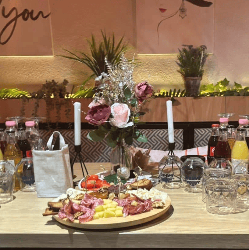 A table with food and drinks arranged: a plate of appetizers, flowers in a glass vase, a candle, and various bottles of drinks. The setting is cozy with decorative elements.