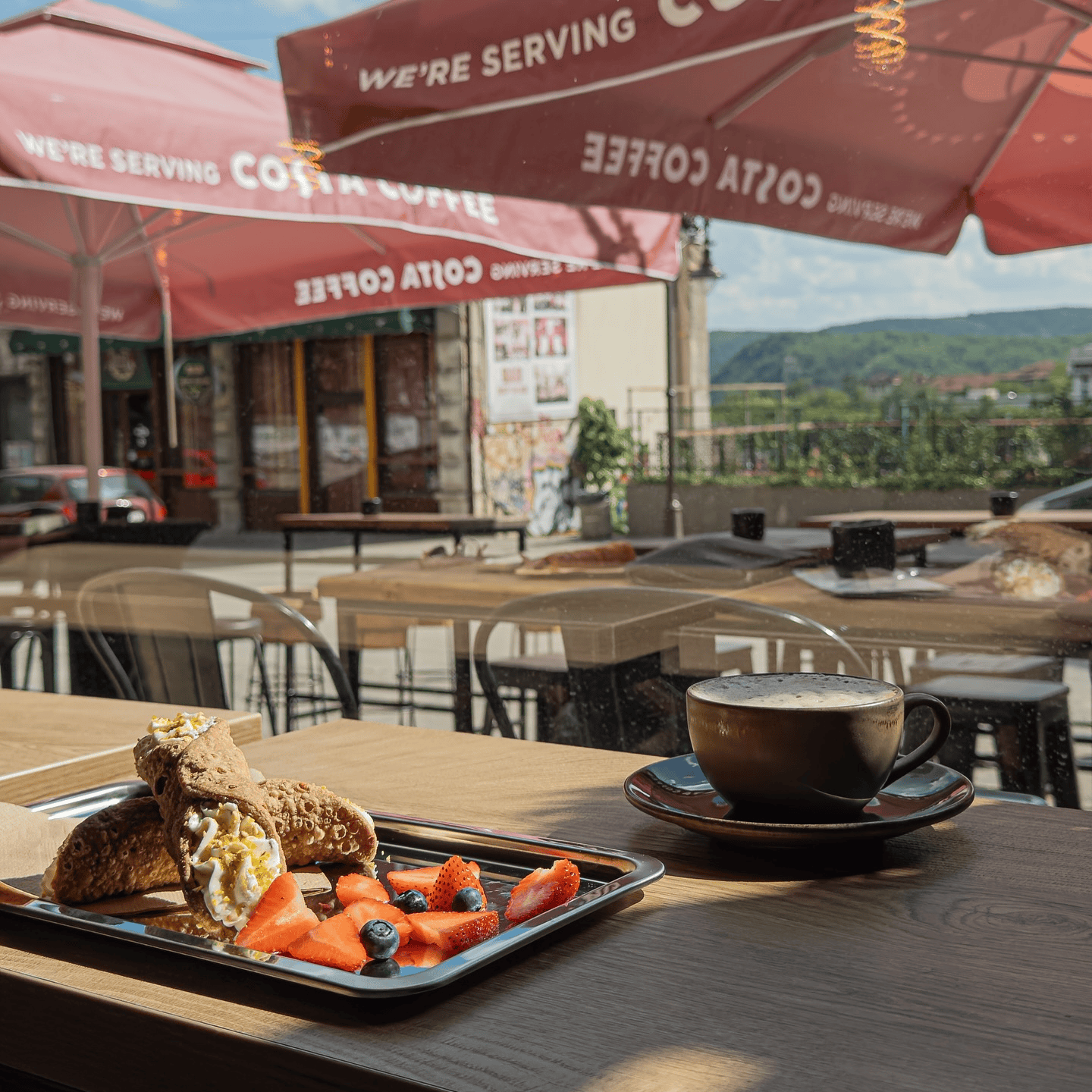 A cup of cappuccino and a plate of desserts (pancakes, strawberries, and blueberries) on a table with a wooden surface. In the background, canopies of a restaurant with signs and greenery are visible.