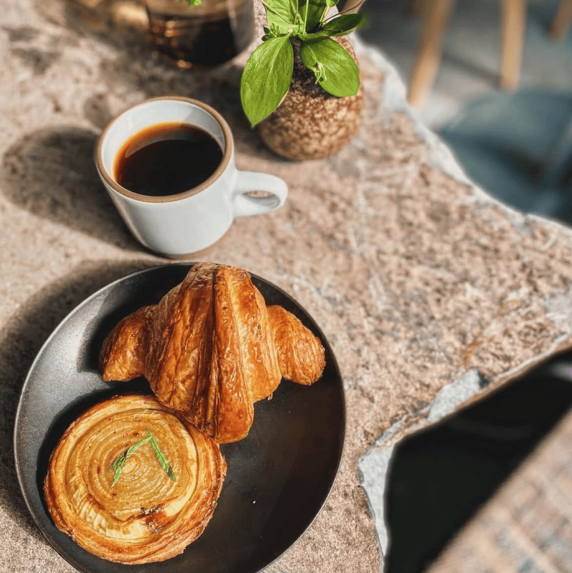 A cup of coffee in a white cup and a plate with two pastries (a croissant and a cake) on a dark background. A potted plant is placed on the table.